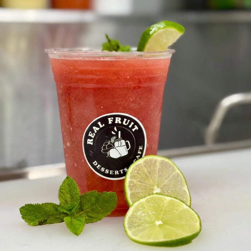 Red fruit drink in a branded cup, garnished with lime wedges and fresh mint on a counter.
