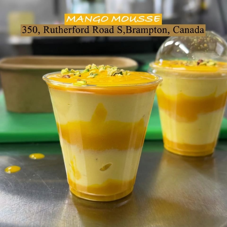 Two layered mango mousse desserts in clear cups, topped with crushed pistachios, displayed on a restaurant kitchen counter.